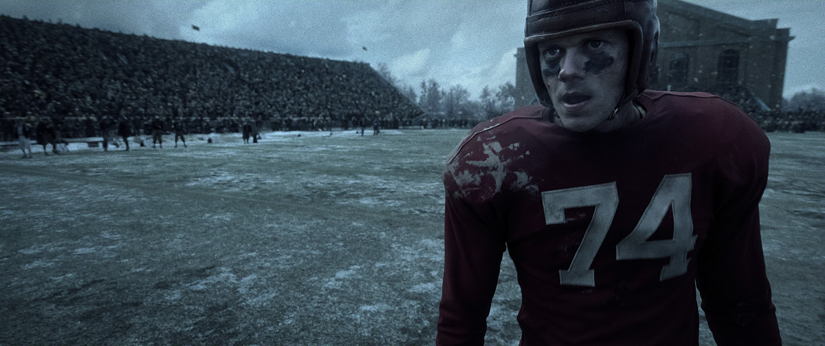 An actor wears a replica of a 1940's Wisconsin football uniform that displays the number 74. Behind him is the recreated Field House on UW–Madison's campus.