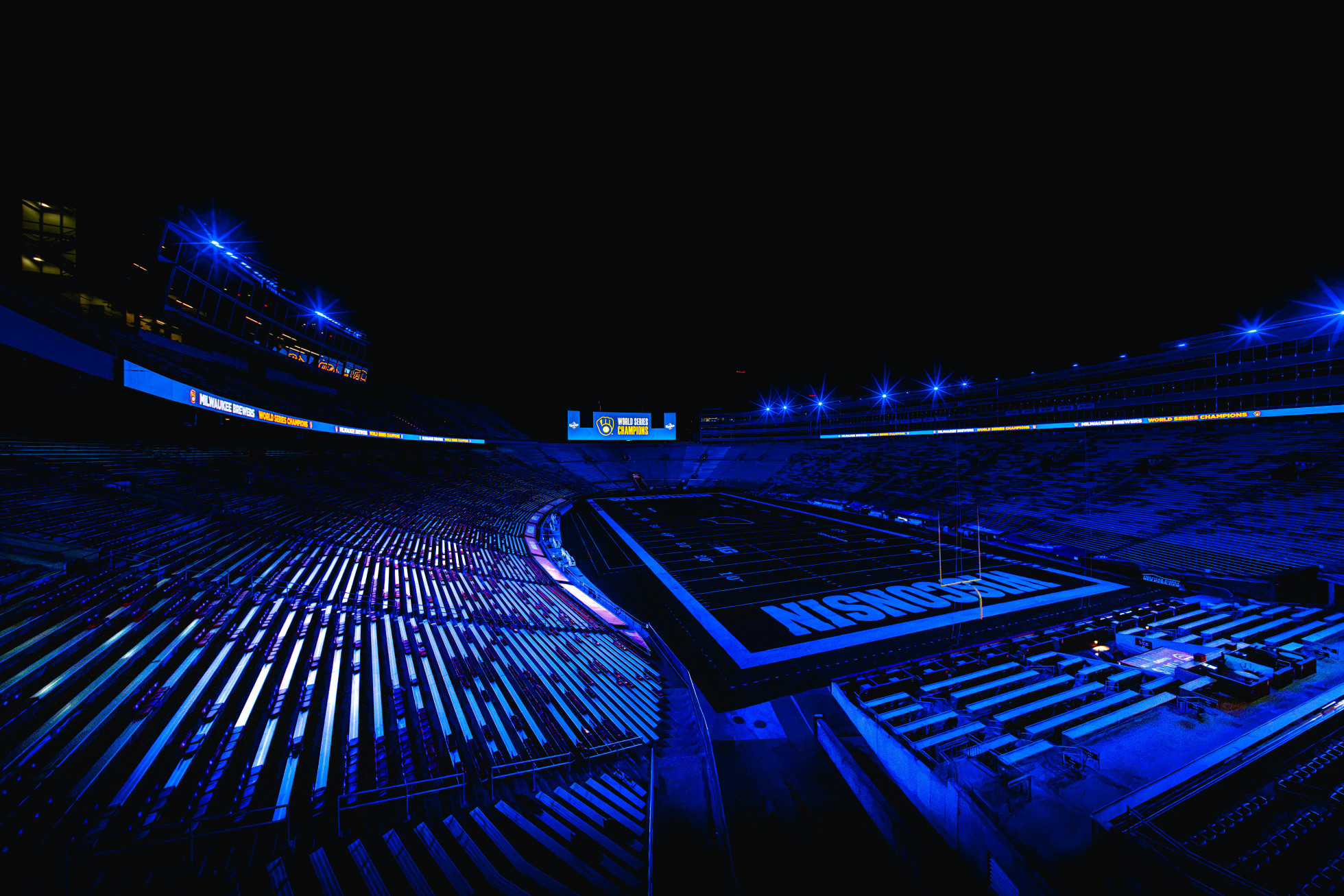 A dimly lit Camp Randall Stadium is shown in full view under blue lights, with the scoreboard showing the Milwaukee Brewers logo and the words "World Series Champions."