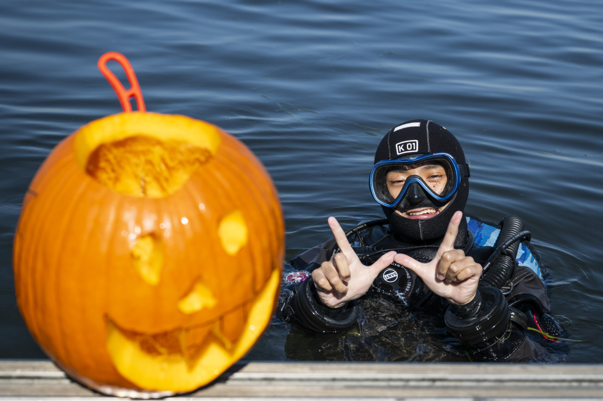 A student wearing SCUBA gear floats in the water and holds up a W sign with his hands. His carved pumpkin rests on the pier in front of him.