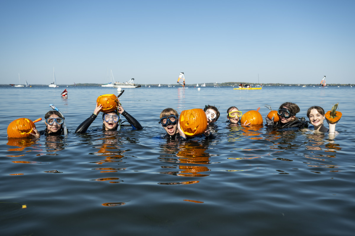 A group of students all wearing SCUBA goggles float in the water and hold up their carved pumpkins for a picture.