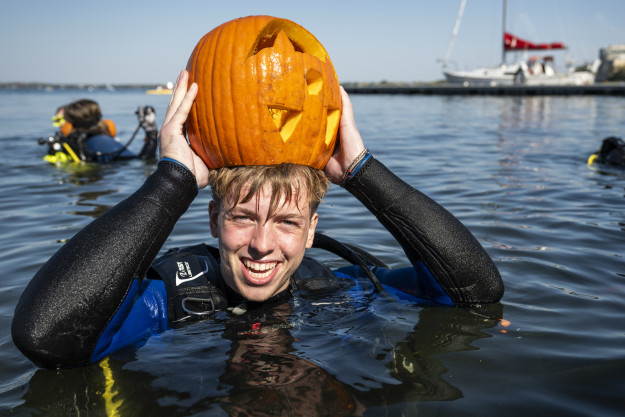 A student floats in the water and wears his carved pumpkin over his head like a hat.