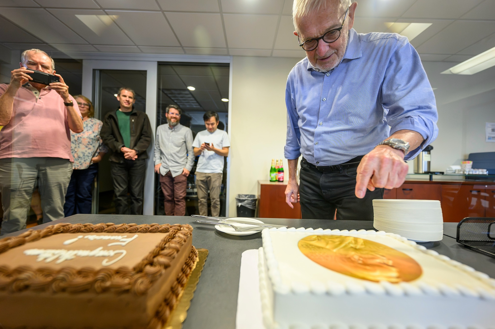 Francis Halzen cuts into a cake with a Nobel Prize medallian design, with colleagues watching in the background.