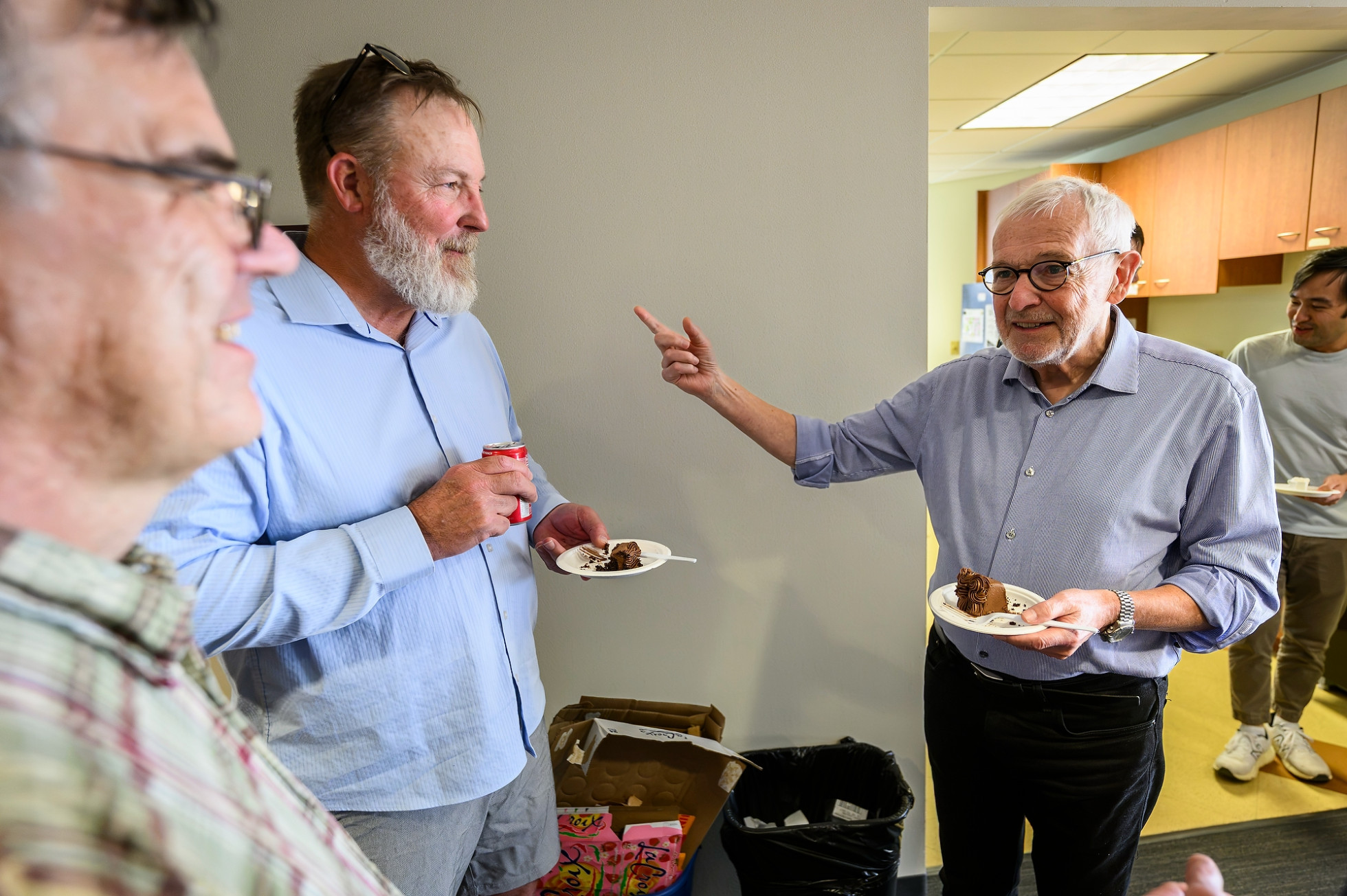 Francis Halzen, holding a piece of cake and pointing with his hand, talks with two colleagues in the office.