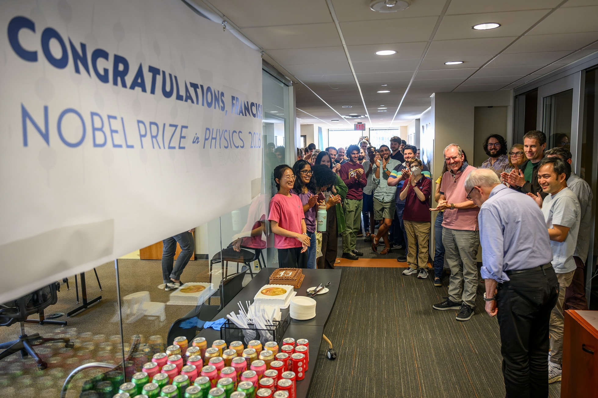 Francis Halzen bows in acknowledgement as his office colleagues greet him in the office hallway, with a table for cake and beverages alongside them. 