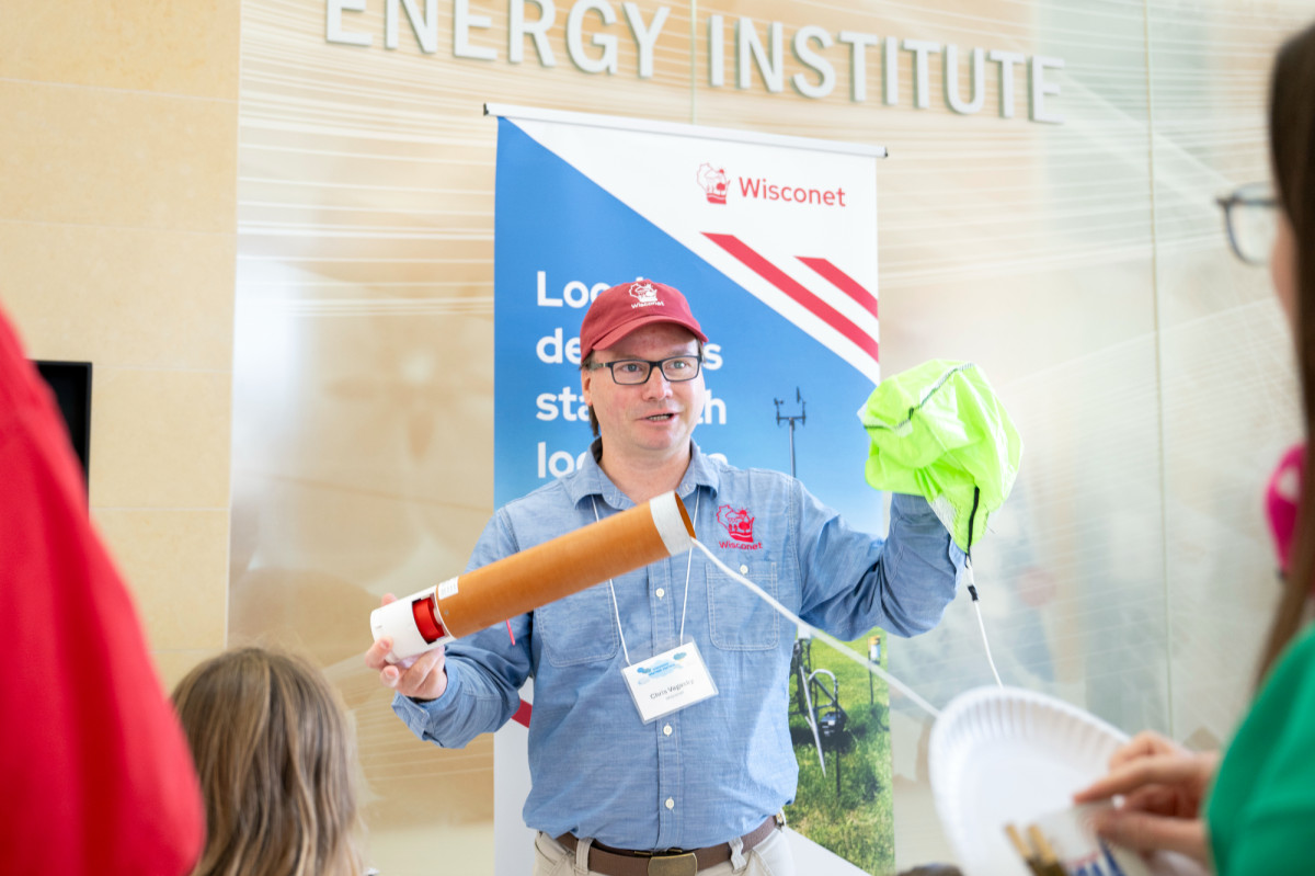 Vagasky wears a red hat displaying the Wisconet logo while showing how a weather balloon works to a nearby crowd of people.