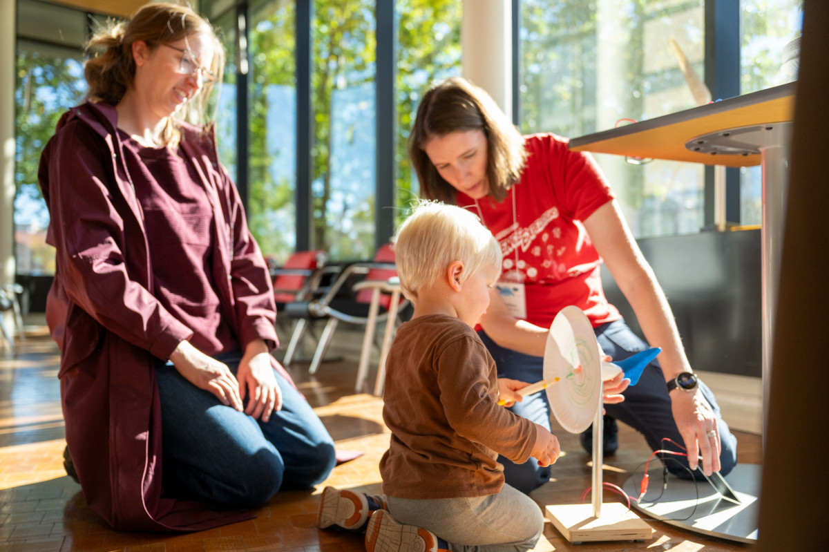 A young child sits on the floor and paints a spinning paper plate operated by a nearby UW faculty member while his mom looks on.