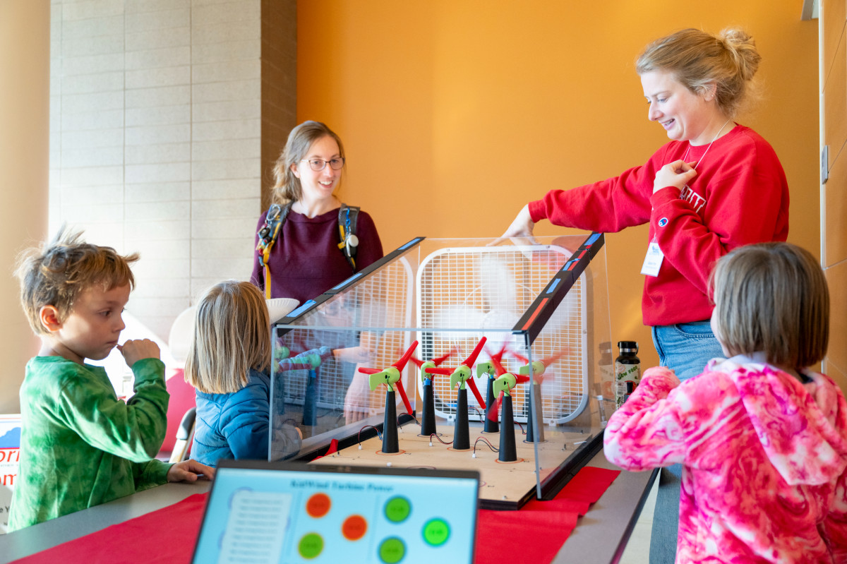 Yum operates a box fan that makes mini wind turbine models spin. Young kids stand nearby watching the demonstration.
