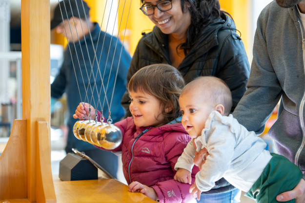 Two parents hold their young children up to a large model of a Newton's cradle device.