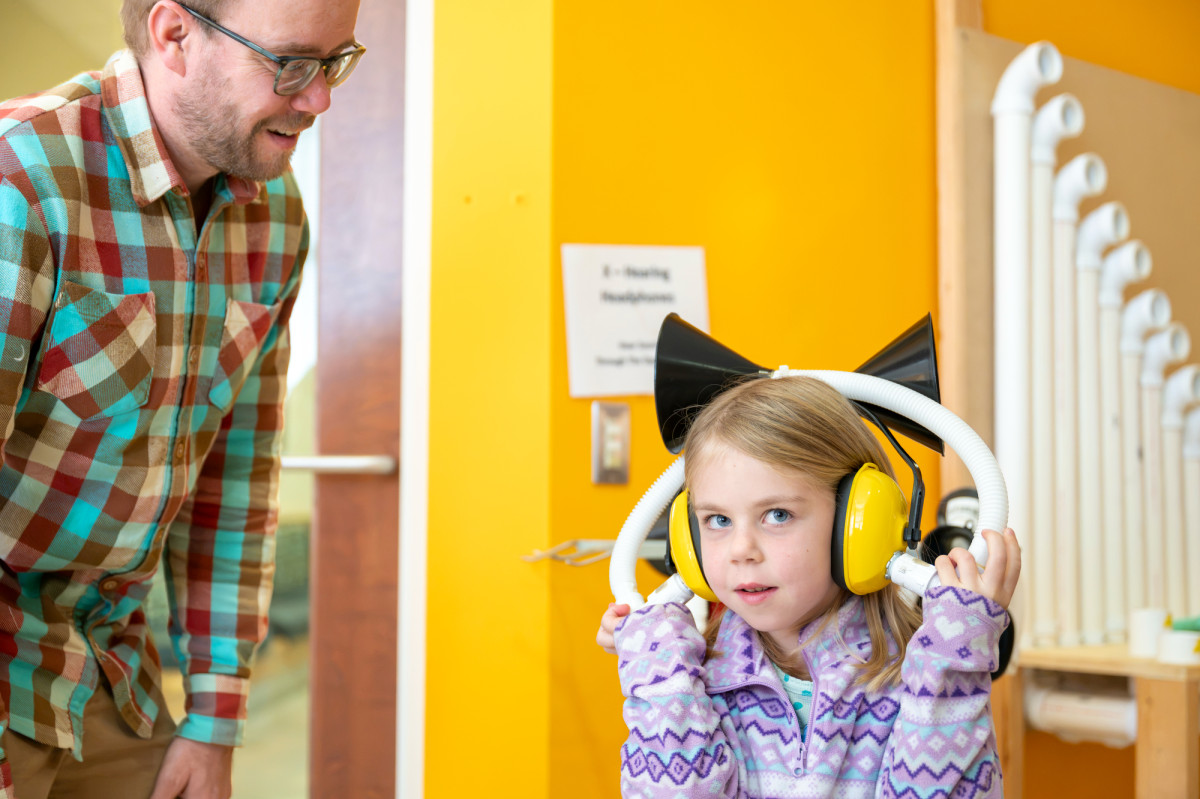 A young girl listens to the sounds around her while wearing a reverse hearing contraption made from a pair of protective ear muffs, tubes and plastic funnels.